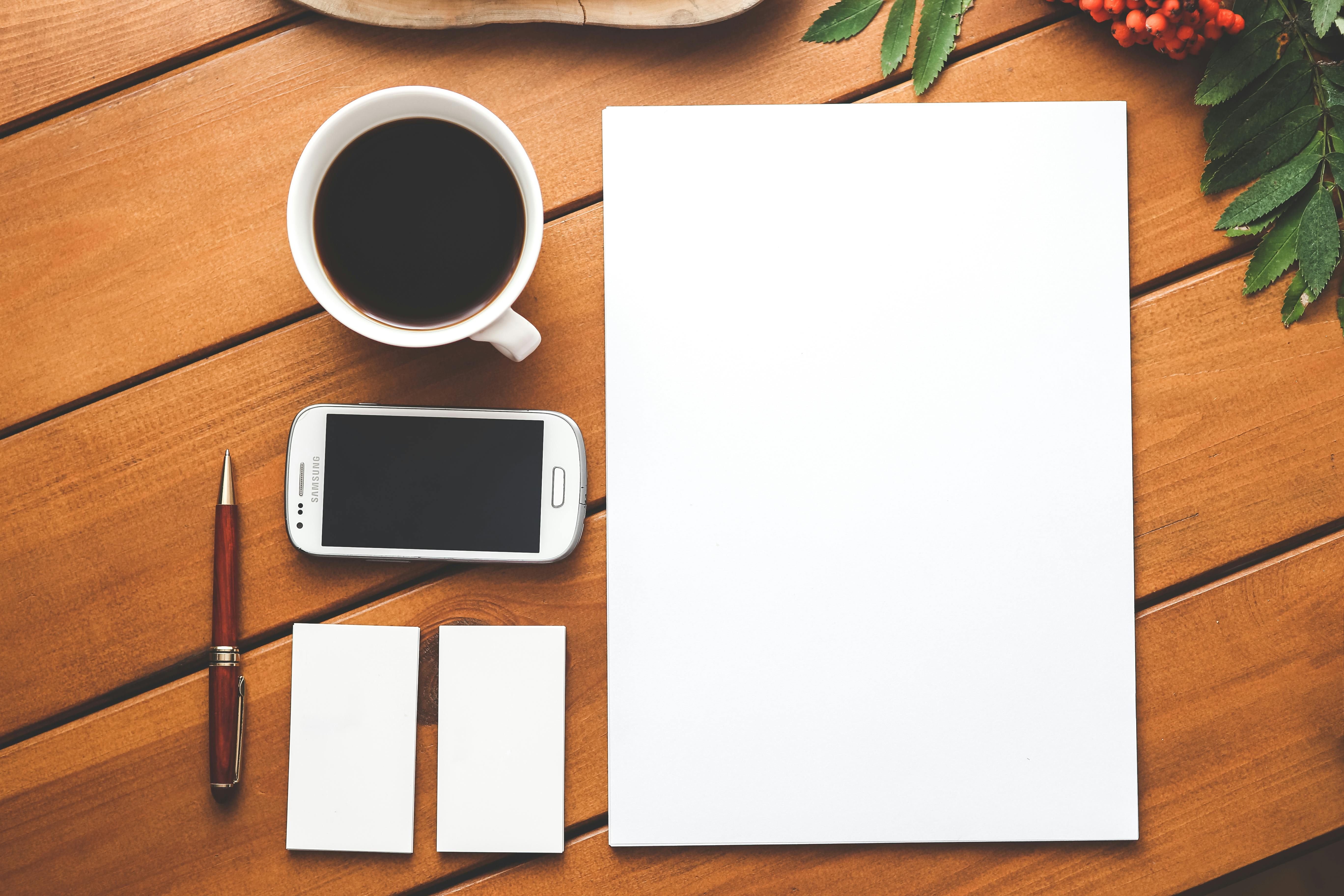 A flat lay of a wooden table with a cup of black coffee, a Samsung smartphone, a red and silver pen, two blank business cards, and a blank sheet of paper. Green leaves and red berries frame the top right corner of the image.
