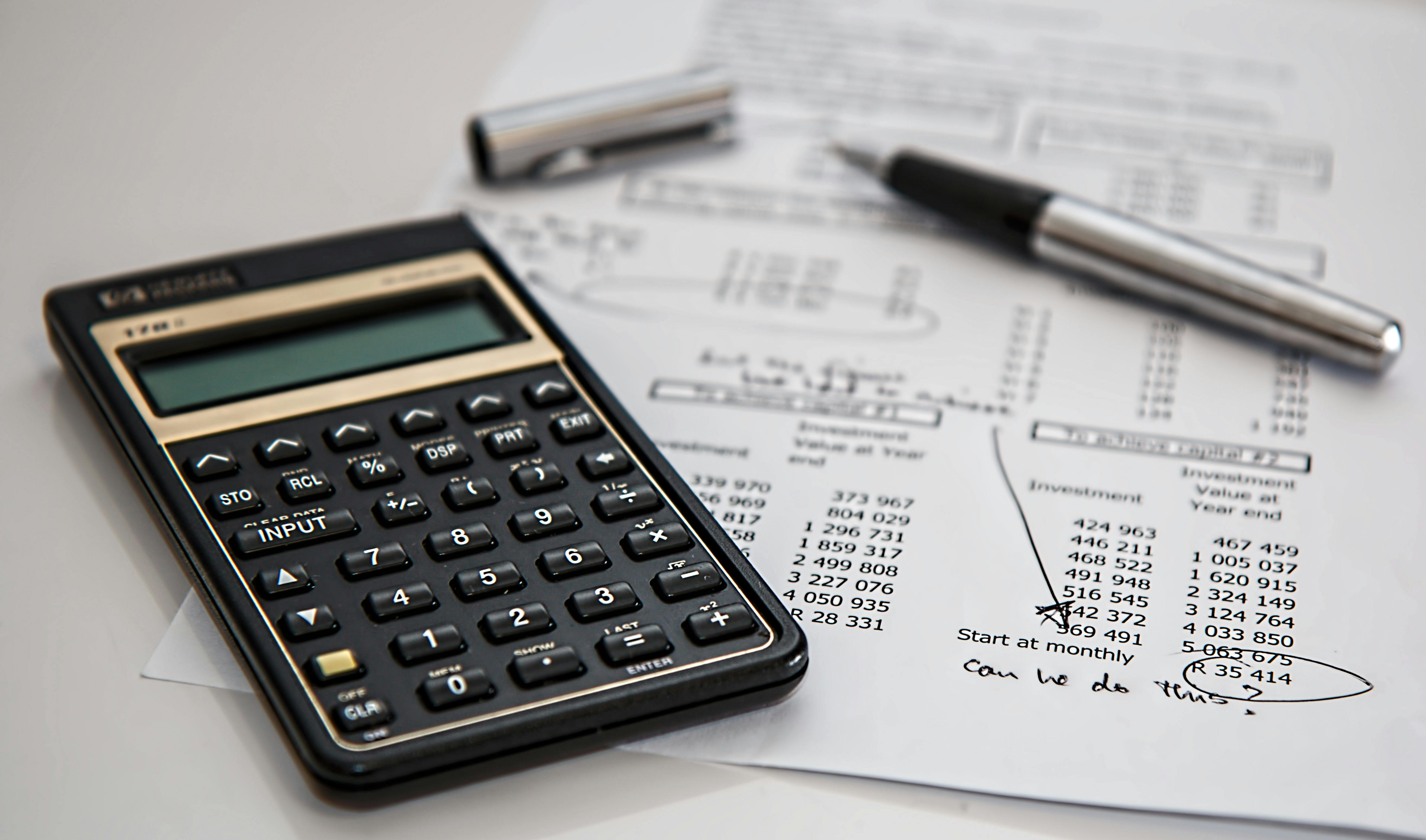 A close-up of a black calculator, a pen, and financial documents with printed numbers and handwritten notes. The paper includes a circled figure and the note 'Can we do this?' written in cursive.